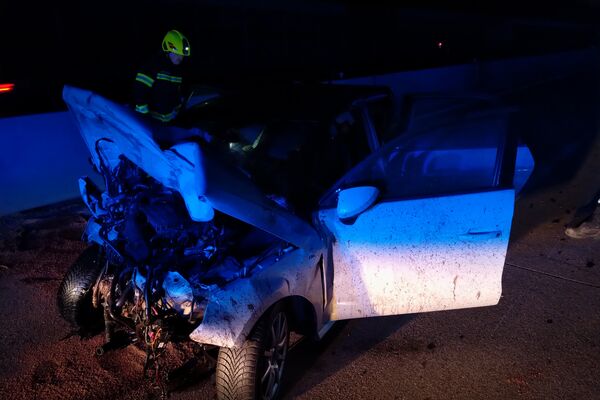 Ein schwer beschädigtes Auto steht nachts auf der Straße, während ein Feuerwehrmann die Unfallstelle untersucht.