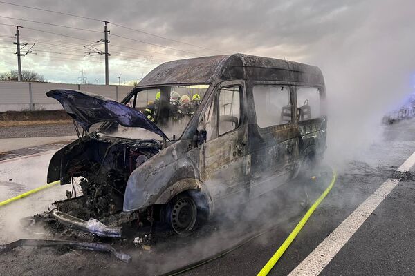 Ein ausgebranntes Fahrzeug steht auf der Straße, während Feuerwehrleute den Brand löschen. | © Name