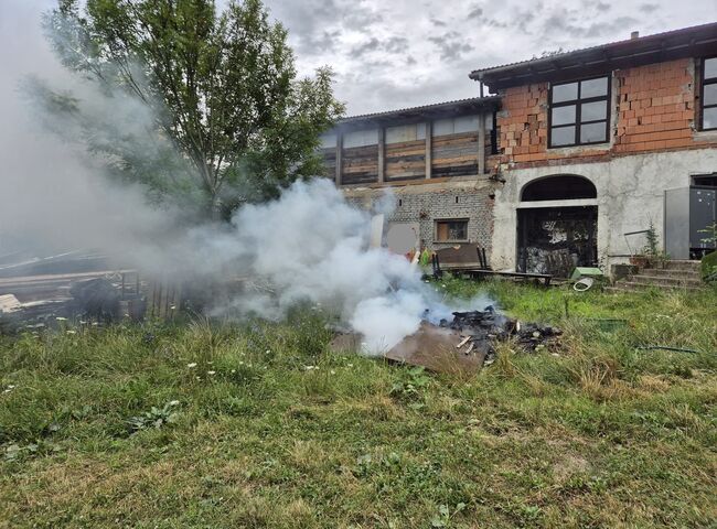 Rauch steigt aus brennendem Holz in einem Garten hinter einem alten, teilweise renovierten Gebäude auf.