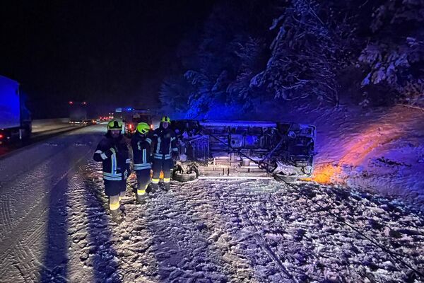 Feuerwehrleute untersuchen einen umgekippten Bus auf einer schneebedeckten Straße in der Nacht. | © Name